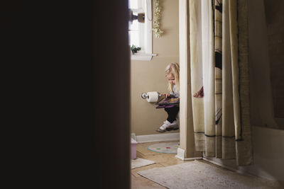 Girl using tablet computer while sitting in bathroom seen through doorway
