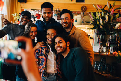 Young woman photographing cheerful multi-ethnic friends standing at restaurant during brunch