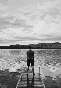 Rear view of man standing on pier in lake against cloudy sky