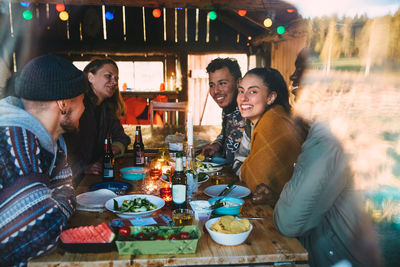 Male and female friends talking while enjoying meal on table in cottage seen through window