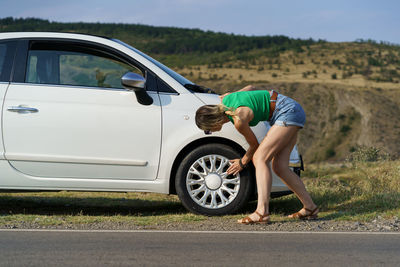 Woman driver parked white car on roadside of highway to check wheel tire on sunny summer day