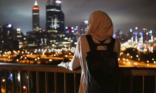 Woman standing by railing against illuminated buildings in city at night