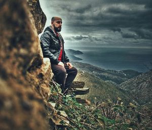 Young man sitting on rock by landscape against sky