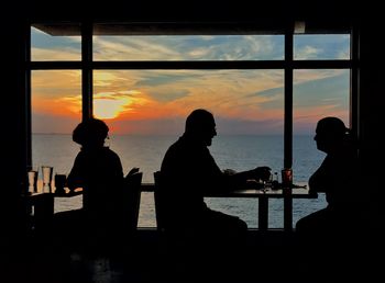 Silhouette people sitting by sea against sky during sunset