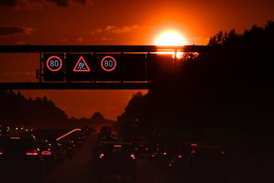 Illuminated road sign against sky at sunset