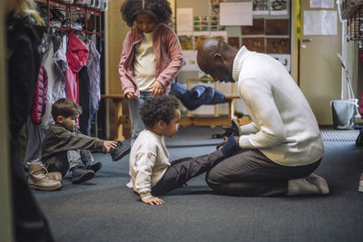 Side view of male teacher helping boy while wearing shoe at kindergarten