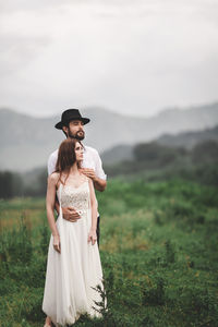 Young couple looking away while standing on grassy field against sky