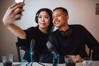 Female blogger taking selfie with male colleague sitting against wall at home