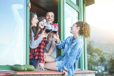 Smiling friends toasting drinks while sitting on window sill against sky