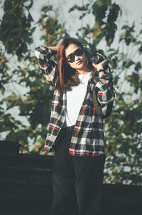 Young woman wearing sunglasses standing against plants is holding slr camera