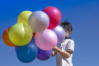 Low angle view of boy holding multi colored balloons against clear blue sky