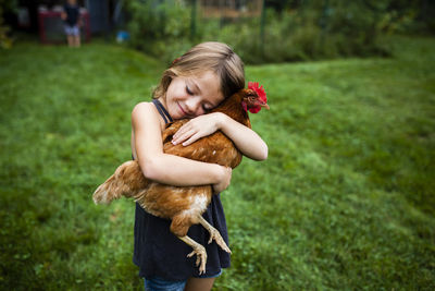 Smiling girl with eyes closed holding hen while standing in yard