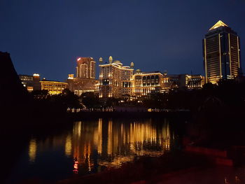 Illuminated buildings by river against clear sky at night