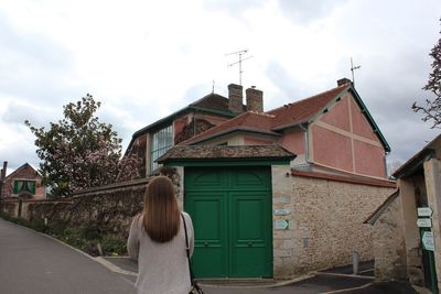 Rear view of woman standing on street by house against cloudy sky