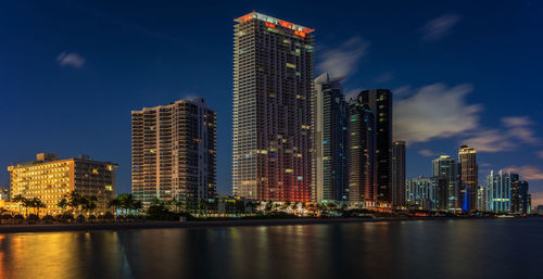 Illuminated city by river and buildings against sky