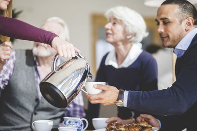 Cropped hand of woman pouring drink in cup held by father while grandparents sitting at table in nursing home