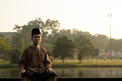 Portrait of man wearing traditional clothing while sitting against lake during sunset