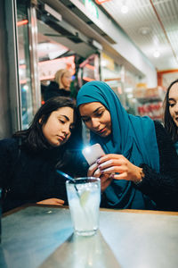 Young muslim woman photographing drink through smart phone while sitting amidst female friends in cafe