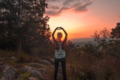 Rear view of woman making heart shape against sky during sunset