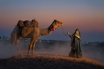 Woman with camel on sand in desert against sky during sunset