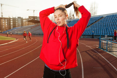 Portrait of woman listening music while holding hair on running track against sky