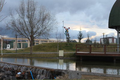 Bare trees and buildings by lake against sky