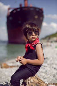 Fashionable boy child with long hair sit on a log next to a large ship that ran aground 