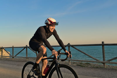 Side view of young man riding bicycle against sea during sunset
