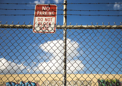 Close-up of information sign hanging chainlink fence against sky