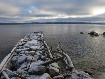 Snow covered groyne over lake against cloudy sky