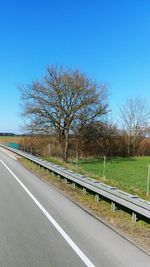 Empty road by bare trees on field against clear blue sky