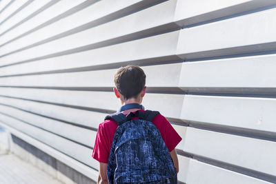 Rear view of schoolboy with backpack walking by wall
