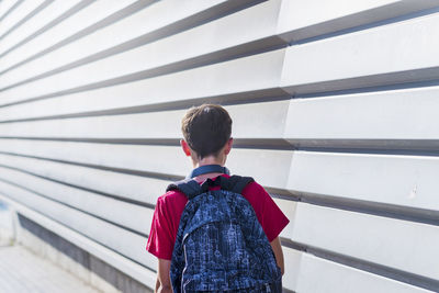 Rear view of schoolboy with backpack walking by wall