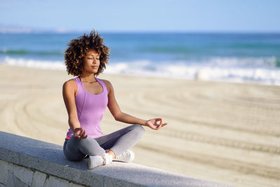 Full length of woman meditating while sitting on retaining wall at beach