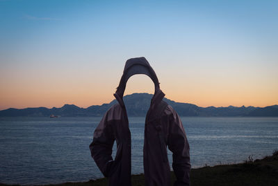 Digital composite image of invisible person wearing hooded jacket while standing by sea against clear sky during sunset