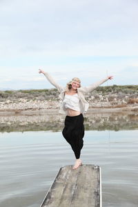 Smiling young woman with arms outstretched while jumping on jetty over lake against sky