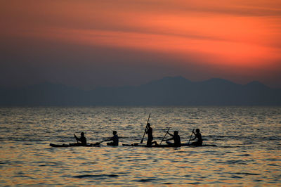 Silhouette people paddleboarding in sea against orange sky