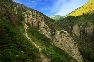 Clay rocks on green mountain slopes with path in summer, sky with clouds