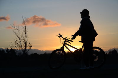 Silhouette man with bicycle against orange sky