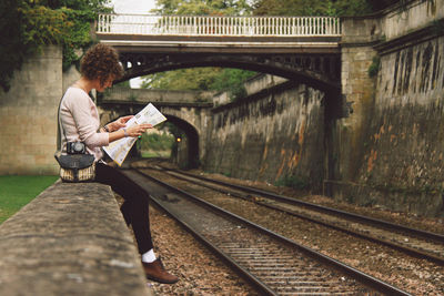 Woman sitting on retaining wall reading newspaper by railroad tracks