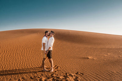 Side view of gay couple kissing while standing at desert against blue sky