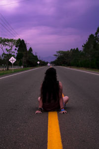 Rear view of woman sitting on road against cloudy sky