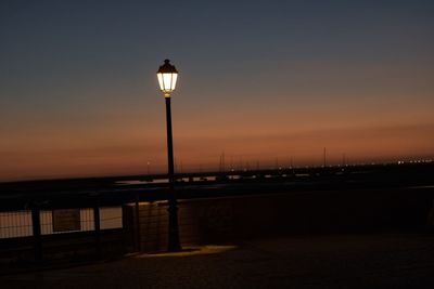 Illuminated street light by sea against sky at sunset