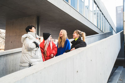 Group of female teen hipsters in stylish casual warm clothing laughing and talking while standing on stairs of modern building