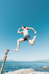 Full length of young woman jumping at beach against clear blue sky