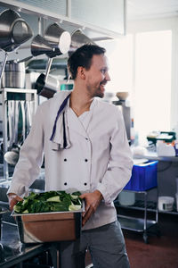 Young male chef smiling while holding container of leaf vegetable in kitchen