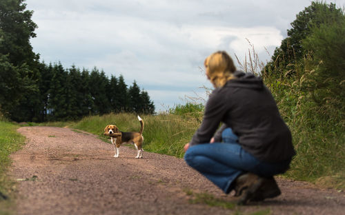 Woman crouching against beagle on dirt road against sky
