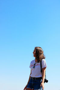 Low angle view of woman with camera standing against clear blue sky