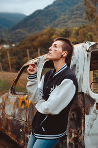 Young female with short hair smoking cigarette while resting near aged burnt vehicle in countryside