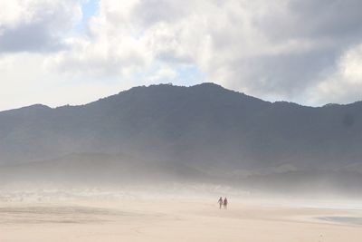 People at beach by mountain against cloudy sky